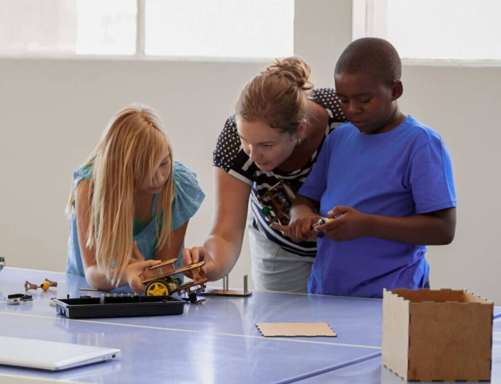 A group of children and an adult gather around a table, working together on robotics projects in a bright classroom.