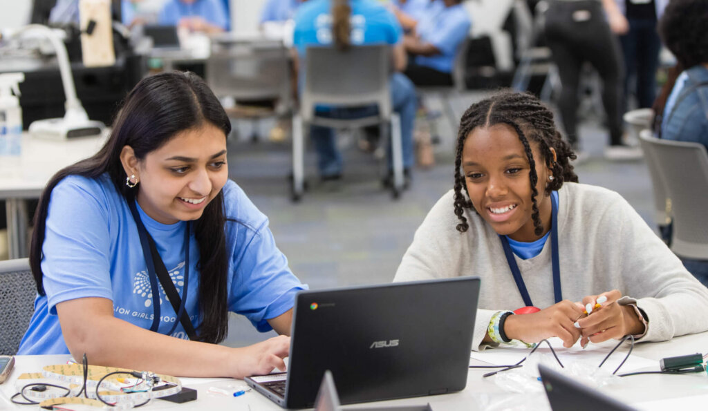 Two young women sit at a table, smiling and working on a laptop together as they collaborate on an AI Challenge in a busy classroom filled with others engaged in group activities in the background.