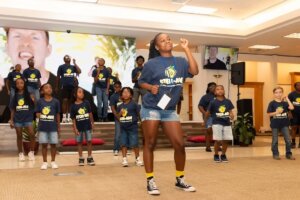 A group of children and adults wearing matching blue shirts and name tags perform a choreographed dance on a stage indoors, with a woman leading in front and a large screen displaying faces behind them—a lively celebration highlighting the spirit of rural workforce development.