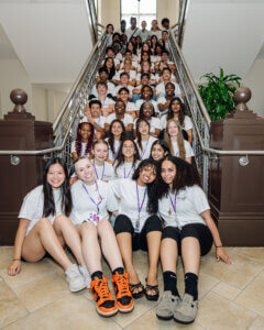 A large group of diverse teens in matching white shirts sits on a staircase, smiling at the camera. Four girls sit in front, and the rest are grouped behind them along the steps in a well-lit indoor space, showcasing proud afterschool STEM leadership and amplifying youth STEM voices.