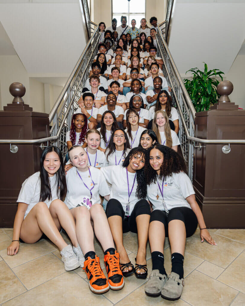 A large group of diverse teens in matching white shirts sits on a staircase, smiling at the camera. Four girls sit in front, and the rest are grouped behind them along the steps in a well-lit indoor space, showcasing proud afterschool STEM leadership and amplifying youth STEM voices.