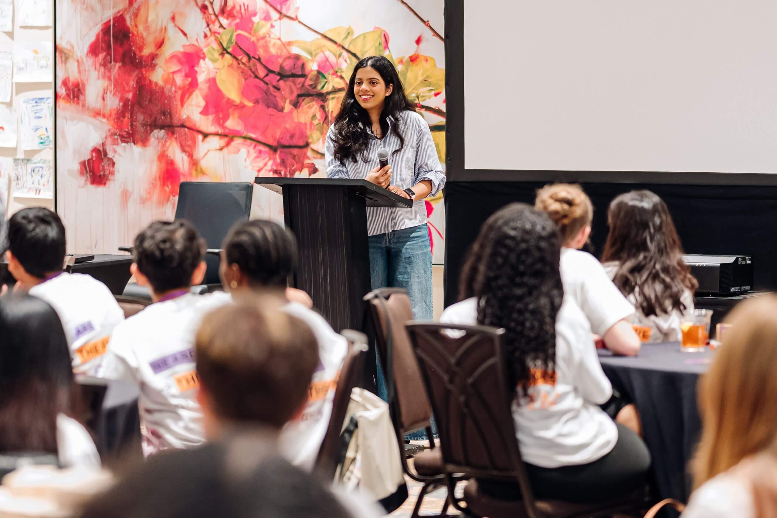 A woman stands at a podium smiling and speaking to a seated audience in a conference room with a colorful floral backdrop behind her, representing the impactful work of the STEM youth ambassador program and highlighting the importance of afterschool STEM leadership.