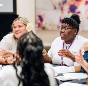 Five people sit around a table, engaged in discussion as part of a STEM youth ambassador program. Papers and pens are on the table. One person in the center speaks, while others listen and smile. The background has a blurred, colorful abstract painting.