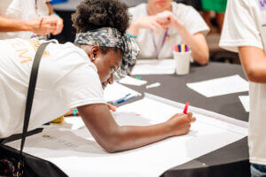 A young person, participating in the afterschool STEM leadership program, leans over a table and writes on a large sheet of paper with a pink marker during a group activity. Others sit nearby, also working, with pens and paper visible on the table.