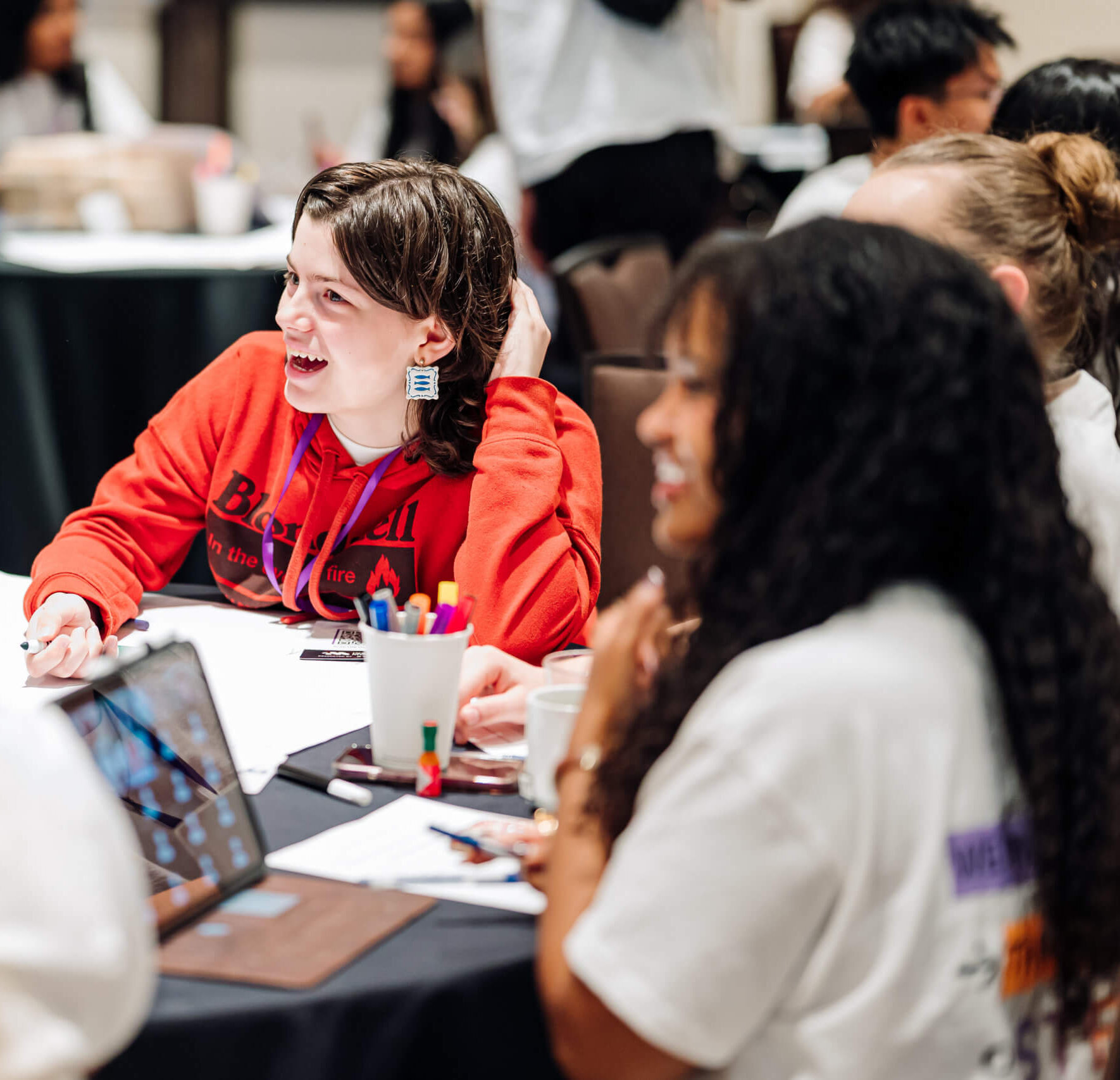 A group of students sits around a table, engaged in discussion as part of an afterschool STEM leadership initiative. One student in a red sweatshirt is smiling and listening. Papers, pens, and a laptop are spread out on the table.