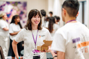 A woman with brown hair, wearing a Flight Crew t-shirt and holding a box, smiles while talking to a man at an indoor event focused on afterschool STEM leadership. Other people are visible in the blurred background.