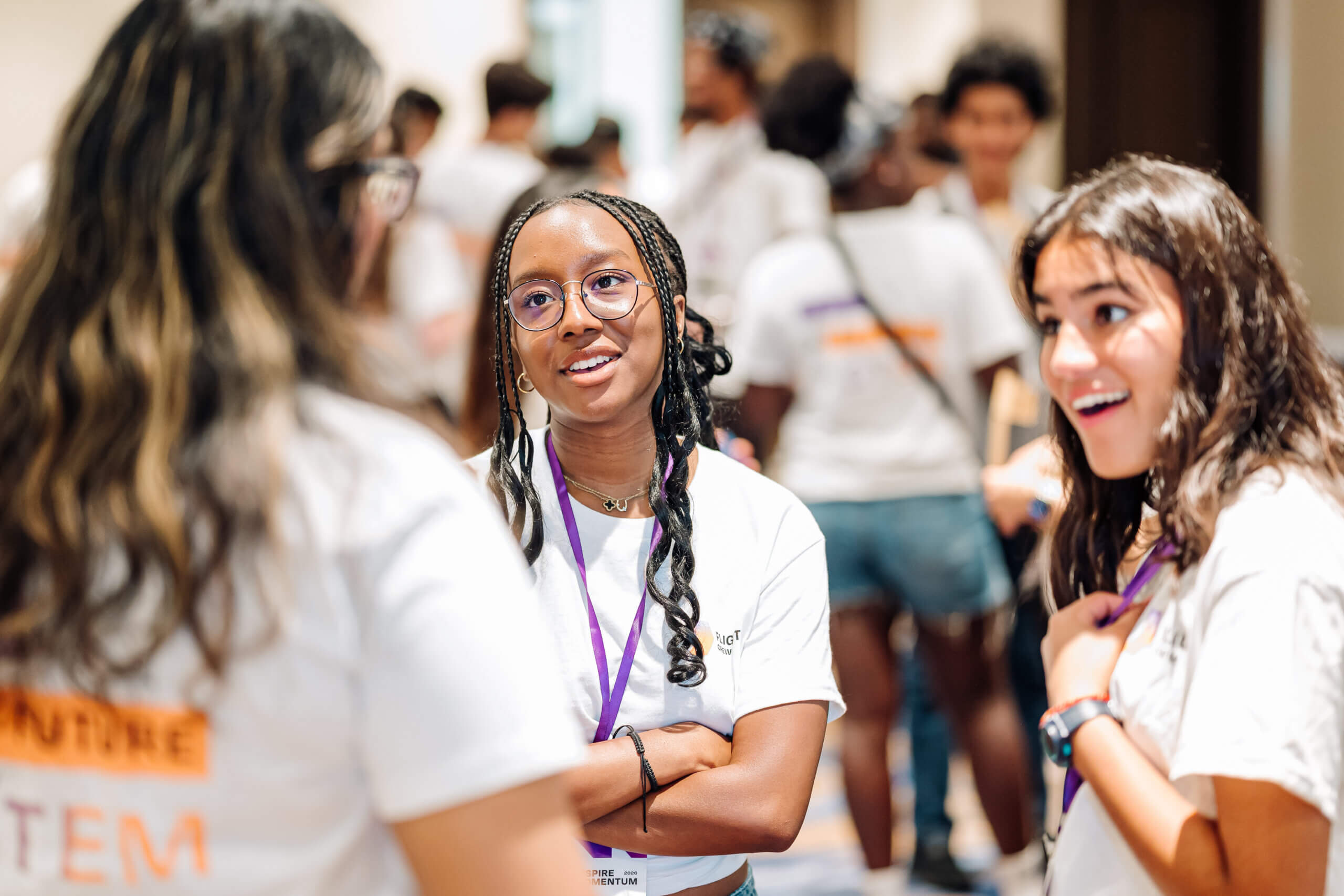 Three young women in white shirts and purple lanyards stand talking and smiling at an indoor event with other people visible in the blurred background, exemplifying youth STEM voices as they engage and connect with fellow participants.