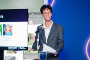 A young man in a gray suit stands at a podium, smiling and holding papers. As a participant in the STEM youth ambassador program, he speaks confidently into the microphone. A screen with photos and text is in the background beside a blue-lit wall, highlighting inspiring youth STEM voices.