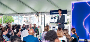 A man in a suit speaks at a podium to an audience seated under a tent, with a screen and lit display behind him, highlighting the impact of afterschool STEM leadership programs on inspiring youth.