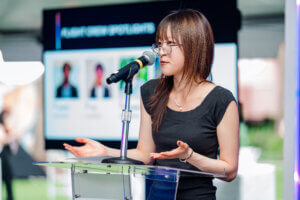 A woman with glasses speaks at a clear podium with a microphone, gesturing with her hands. A blurred presentation screen with photos and text is visible in the background, highlighting the impactful work of the STEM youth ambassador program.
