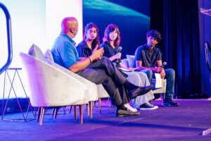 Four people sit on stage chairs having a panel discussion; three hold microphones, listening to the person speaking. A large screen displays an abstract blue and black background behind them. The conversation highlights youth STEM voices, showcasing insights from participants in an afterschool STEM leadership initiative.