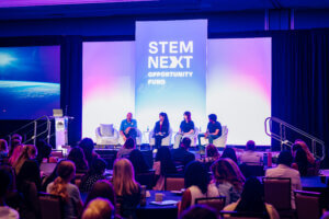 A panel of four people speaks on stage at a STEM NEXT OPPORTUNITY FUND event, highlighting the importance of youth STEM voices and the impact of the STEM youth ambassador program, while facing an audience seated at round tables in a conference room.