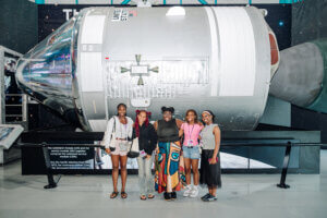 Five young women stand smiling in front of a large space capsule display at a museum, proudly representing the STEM youth ambassador program, with informational signs and a starry background visible behind them.