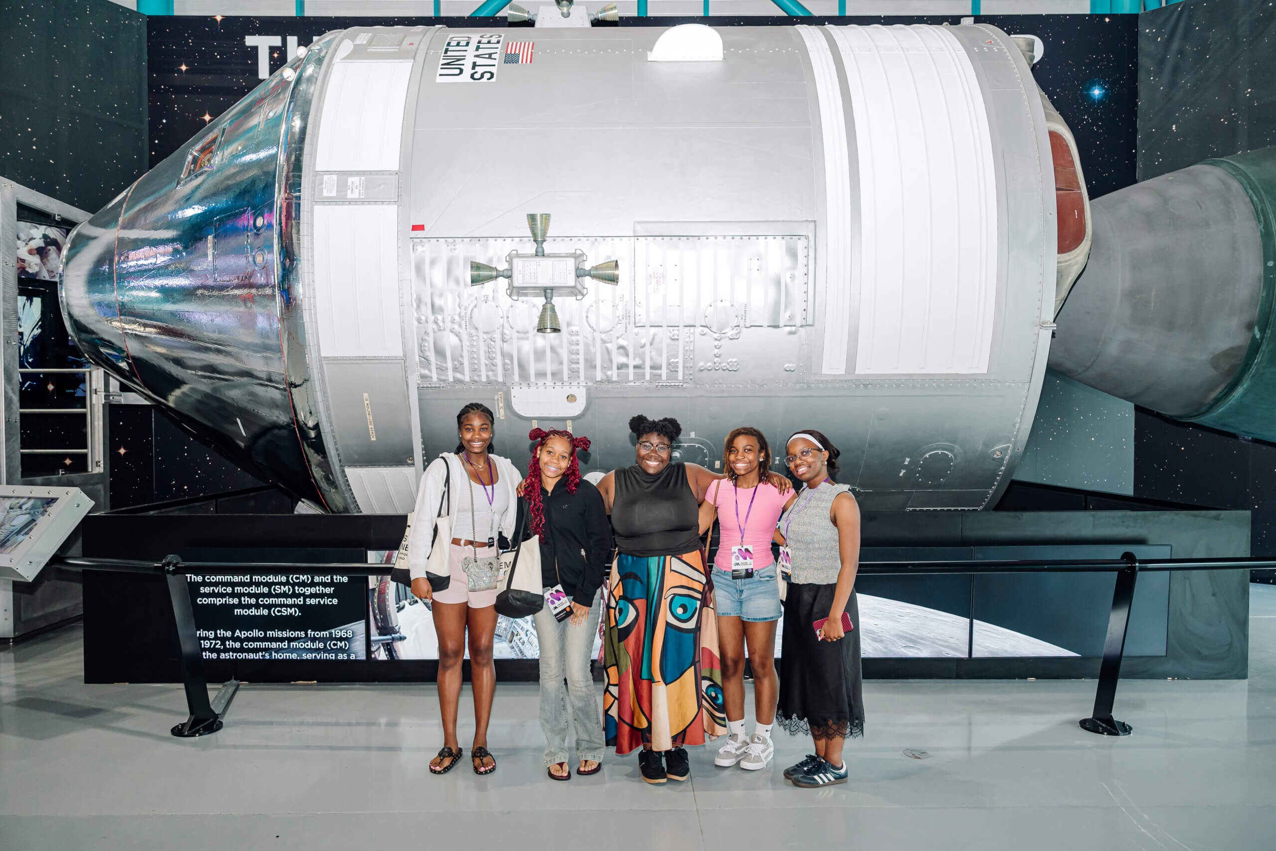 Five young women stand smiling in front of a large space capsule display at a museum, proudly representing the STEM youth ambassador program, with informational signs and a starry background visible behind them.