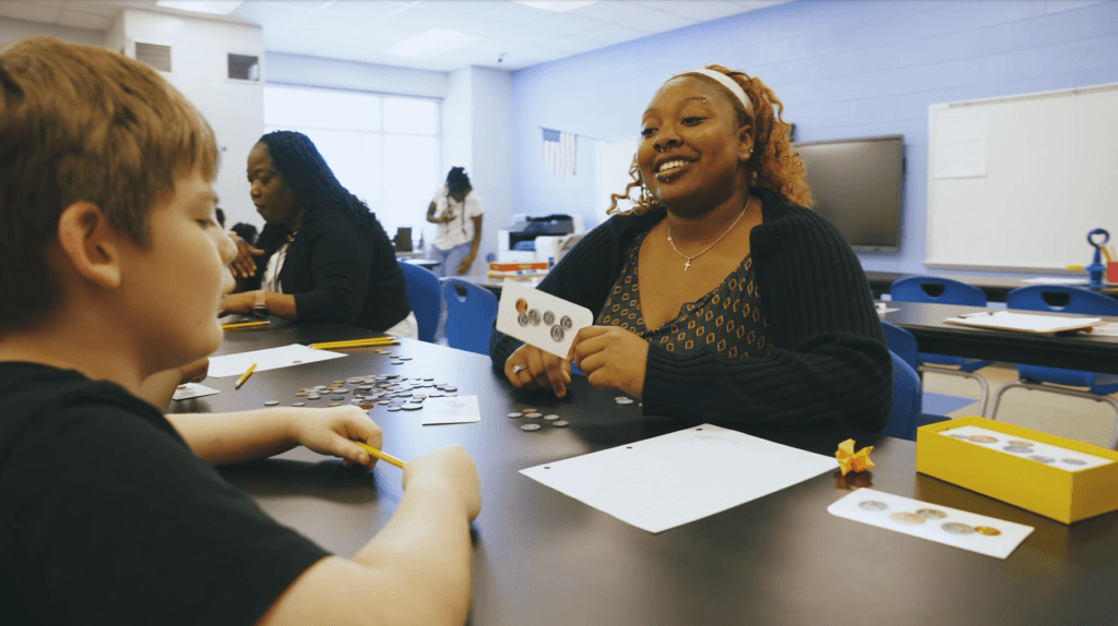 A teacher smiles while holding a card with coin images, sitting at a table with a student in a classroom. Other people work in the background.