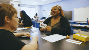 A teacher smiles while holding a card with coin images, sitting at a table with a student in a classroom. Other people work in the background.