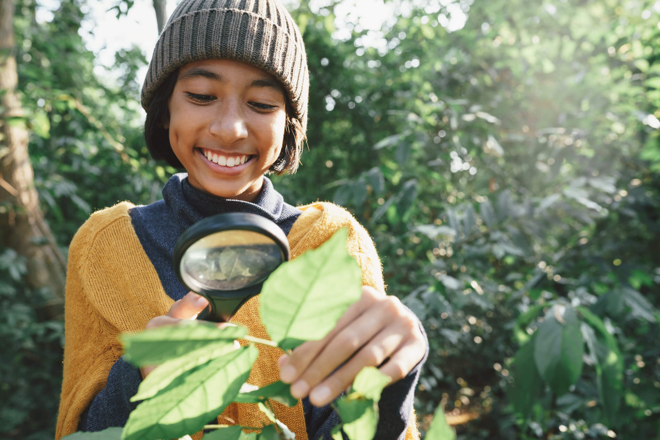 A smiling child wearing a beanie and yellow sweater examines green leaves with a magnifying glass in a sunlit, leafy outdoor setting.