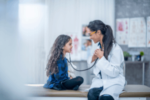 A doctor in a white coat smiles at a young girl sitting on an exam table. The girl holds a stethoscope to the doctor’s chest, exploring Health and life sciences STEM resources in a hands-on way. Medical posters and equipment are visible in the background.
