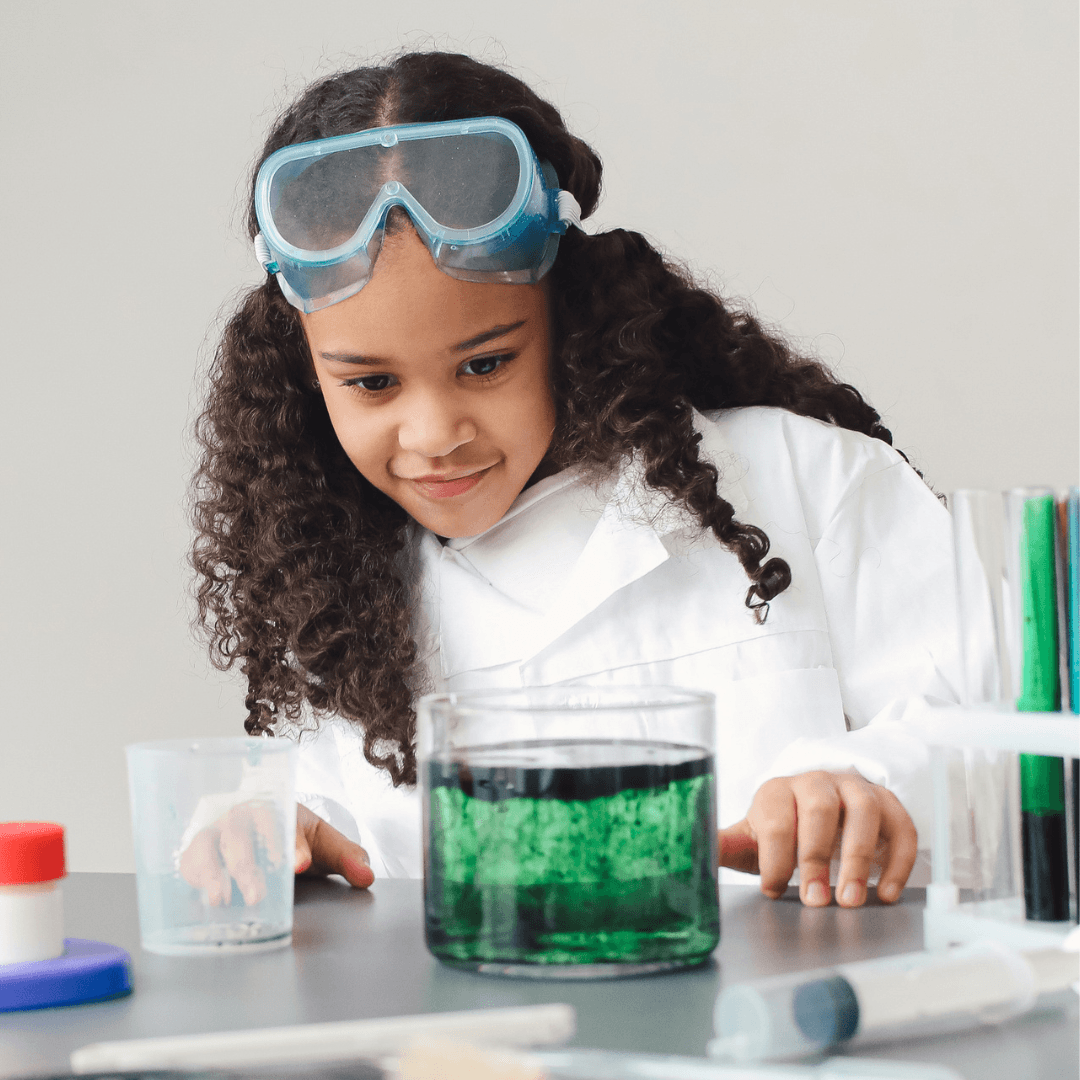 A young girl wearing safety goggles and a lab coat observes a beaker filled with green liquid on a lab table, surrounded by scientific equipment from the Launching Futures Toolkit.