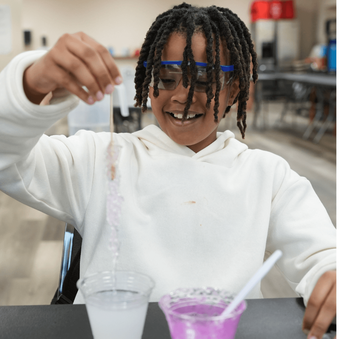 A smiling child wearing blue safety goggles holds up a crystal-covered string over a clear cup, exploring with curiosity—an activity inspired by the Launching Futures Toolkit. Another cup and a spoon sit nearby on the table, ready for more fun experiments.