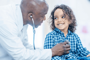 A doctor uses a stethoscope to listen to a smiling child’s chest during a medical exam. The child, wearing a blue checked shirt, looks happy and relaxed while sitting on an exam table—a moment that highlights the importance of health and life sciences STEM resources in supporting children’s well-being.