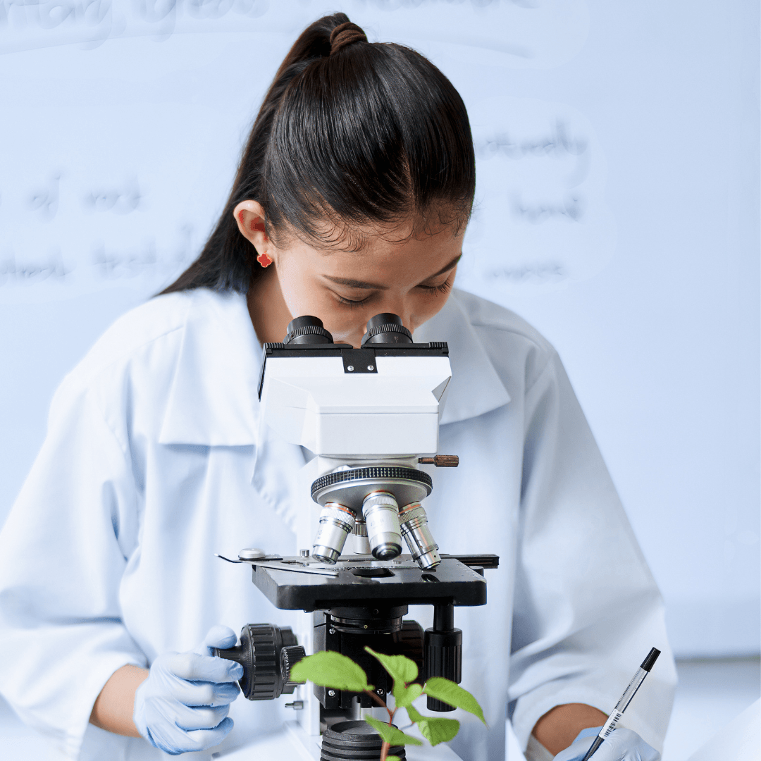 A woman wearing a lab coat and gloves looks into a microscope, examining a green plant sample. She holds a pen, referencing the Launching Futures Toolkit as she works, with a whiteboard and handwritten text blurred in the background.