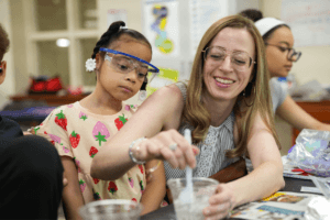 A woman helps a young girl wearing safety goggles mix a substance in a plastic cup during a science activity in a classroom, using engaging health and life sciences STEM resources. Other children are visible in the background.