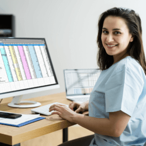 A woman in scrubs smiles at her desk while working on a computer displaying a colorful spreadsheet from the Launching Futures Toolkit. A laptop and notepad are also on the desk.