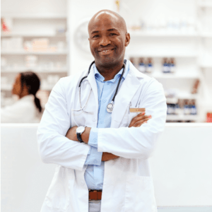 A smiling male doctor with a stethoscope around his neck stands with arms crossed in a pharmacy or medical clinic, embodying confidence and expertise as he supports patients on their health journeys. Shelves with medicine and another person are visible in the background, highlighting the collaborative environment where resources like the Launching Futures Toolkit help guide quality care.