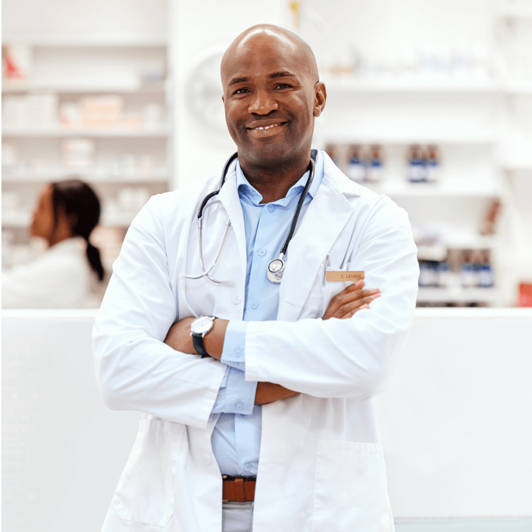 A smiling male doctor with a stethoscope around his neck stands with arms crossed in a pharmacy or medical clinic, embodying confidence and expertise as he supports patients on their health journeys. Shelves with medicine and another person are visible in the background, highlighting the collaborative environment where resources like the Launching Futures Toolkit help guide quality care.