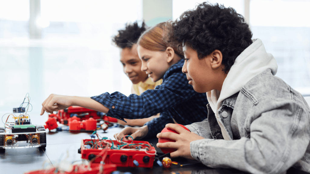 Three children are sitting at a table, engaging with robotics kits and electronic components, appearing focused and curious as they work together on STEM projects.