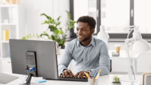 A man in a light grey shirt smiles while typing on a keyboard at a desk in a bright, modern office with plants, lamps, and office supplies around him, thoughtfully engaged with the Launching Futures Toolkit.