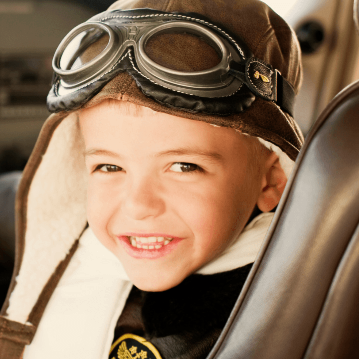 A young child wearing aviator goggles and a brown pilot hat smiles while sitting in an airplane cockpit, with flight controls and instrument panels visible in the background—ready to soar with the Launching Futures Toolkit.