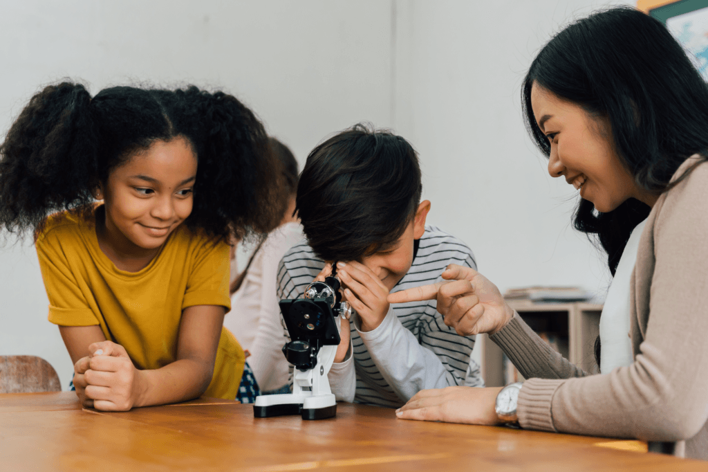 A woman and two children sit at a table. One child looks through a microscope while the others watch and smile. The woman is pointing at the microscope, teaching or explaining something using engaging Health and life sciences STEM resources.