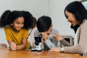 A woman and two children sit at a table. One child looks through a microscope while the others watch and smile. The woman is pointing at the microscope, teaching or explaining something using engaging Health and life sciences STEM resources.