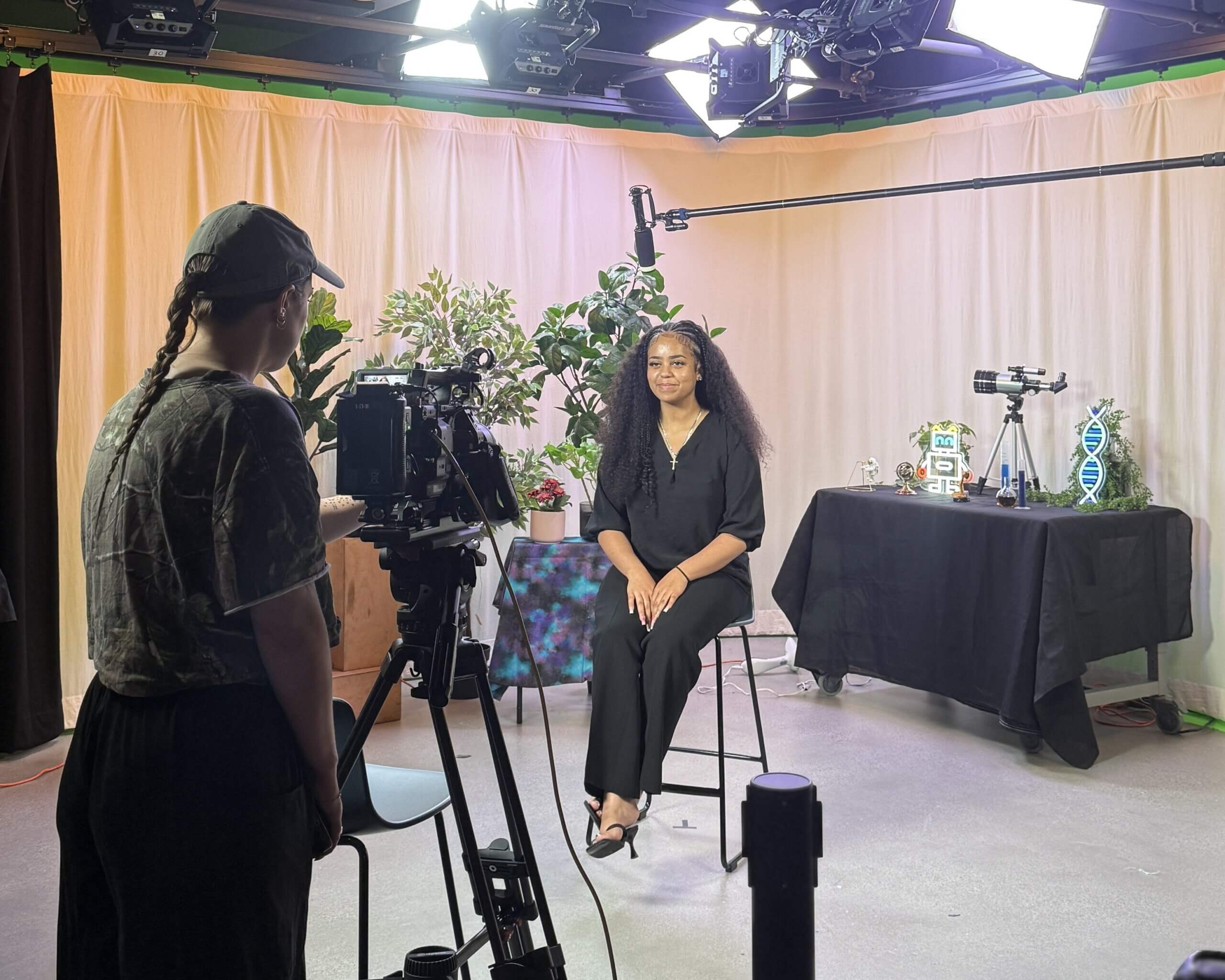 A person sits on a stool in a studio, facing a camera operator. Behind them are plants and a table with science-themed items like models of DNA and a telescope, highlighting the environment of an afterschool STEM leadership initiative. Bright lights illuminate the scene as youth STEM voices prepare to inspire others through engaging storytelling.